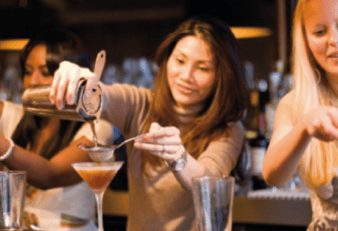 A group of participants in a cocktail making class, gathered around a bar station, mixing ingredients and preparing drinks under the guidance of an instructor.