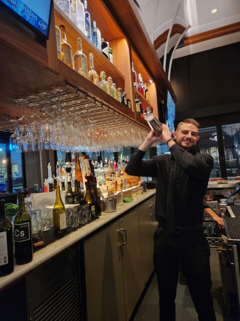 A bartender, Daniel Vinogradov, at Del Frisco's in Fort Lauderdale, smiling while mixing drinks behind the bar.