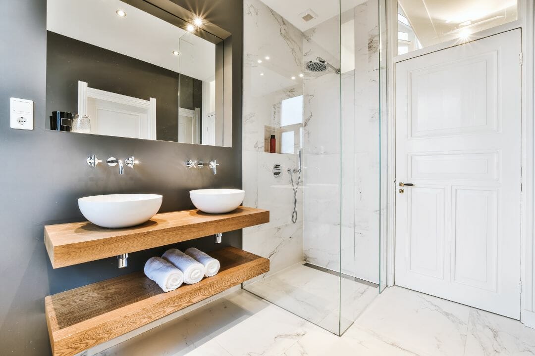 Moderne badkamer met houten wastafel, marmeren muren, glazen douchecabine en witte deur. Bezoek onze winkel voor al uw verbouwwensen.
