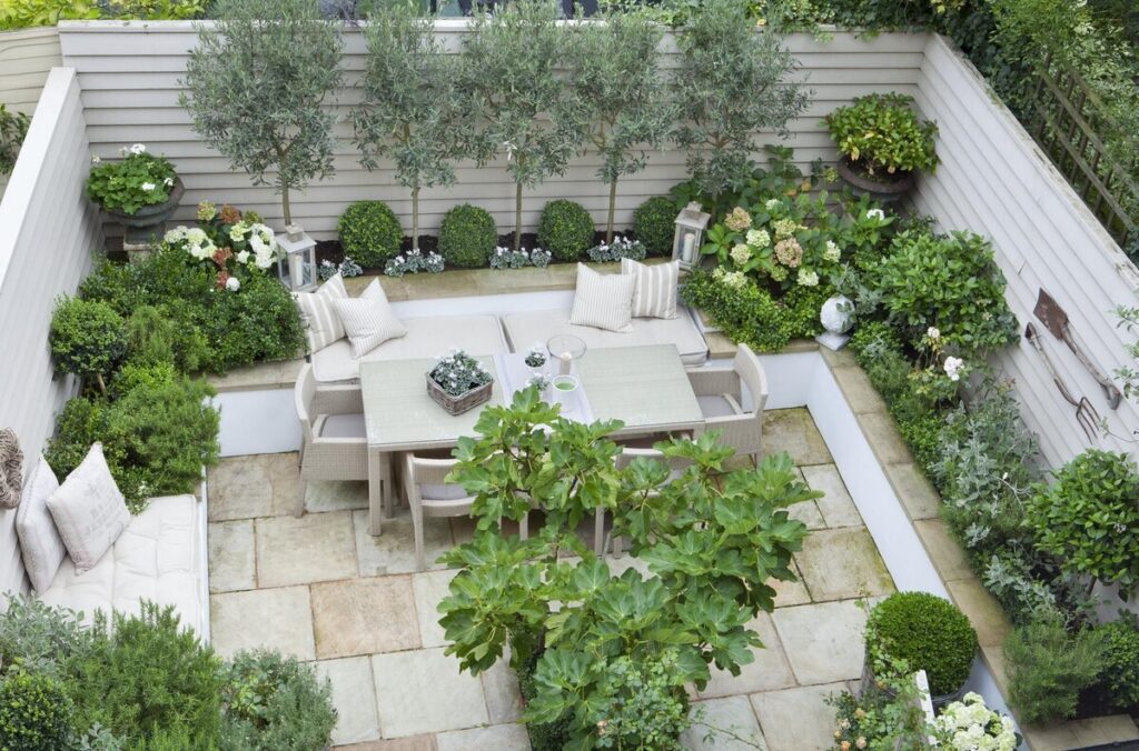 Un petit jardin paysagé en patio, clos, comprenant un ensemble de table à manger entouré d'une végétation luxuriante, incluant des arbustes, des plantes en pot et de petits arbres. Cet espace aménagé par un paysagiste dispose d'un sol en pierre claire et est entouré de clôtures en bois. L'entreprise de paysagisme a conçu cette prestation paysagère pour créer une ambiance apaisante et harmonieuse.