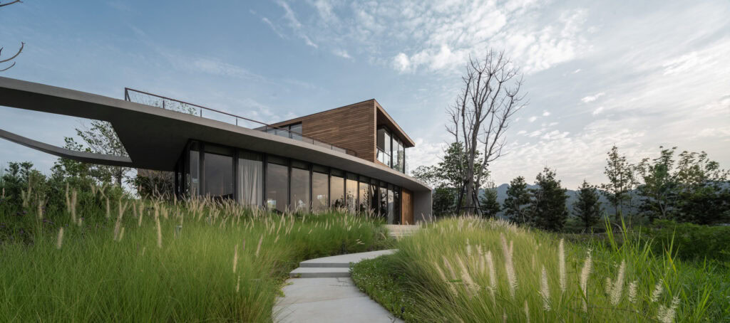 Ce projet se déploie autour d'une villa d'architecte contemporaine, avec de grandes fenêtres et une section supérieure en bois, intégrée dans un environnement où l'architecture rencontre la nature. Les abords sont conçus avec des herbes hautes et des arbres pour créer un cadre naturel et harmonieux. Un chemin en béton aux lignes courbes guide les visiteurs jusqu'à l'entrée principale, combinant esthétisme et fonctionnalité.