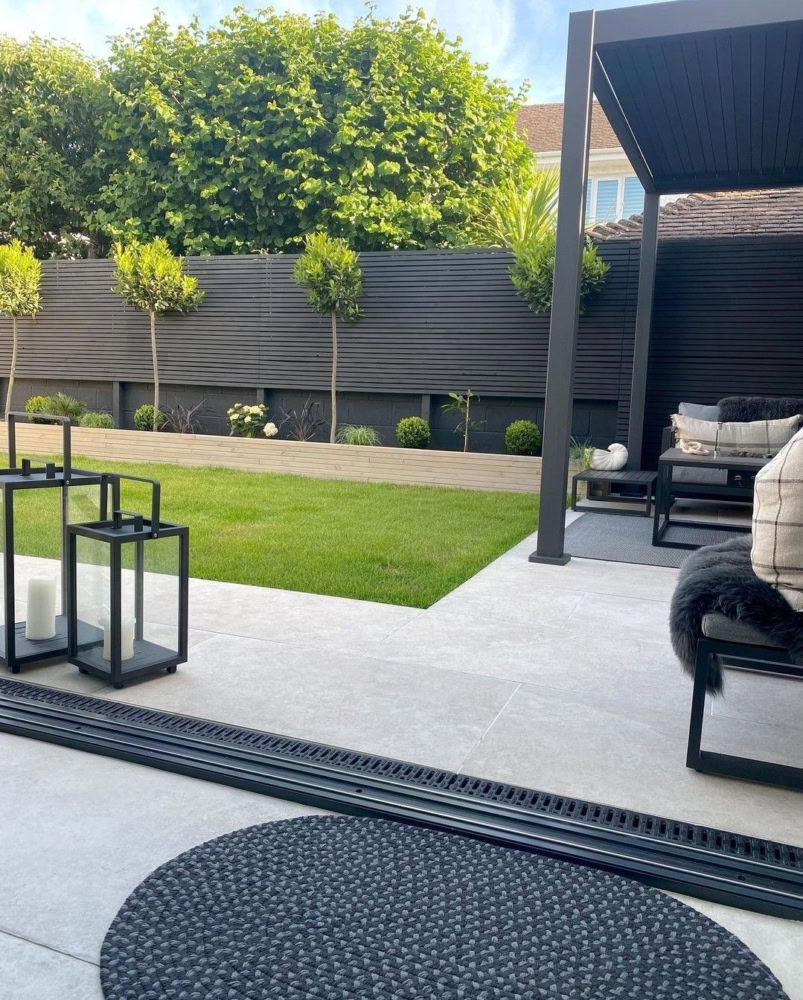 Auf einer modernen Terrasse mit stilvoller Pergola sorgen gepolsterte Sitzgelegenheiten und dekorative Laternen für Charme. Im Hintergrund befindet sich ein angelegter Garten mit gepflegtem Rasen und kleinen Pflanzen, eingerahmt von einem schwarzen Lattenzaun und hohen Hecken. Diese Entwicklungen sind typisch für eine Landschaftsbauleistung, die von einem Landschaftsbauunternehmen oder einem erfahrenen Landschaftsbauunternehmen durchgeführt wird. Die Kunst der Landschaftsgestaltung spiegelt sich in jedem Detail dieses harmonischen Außenbereichs wider.
