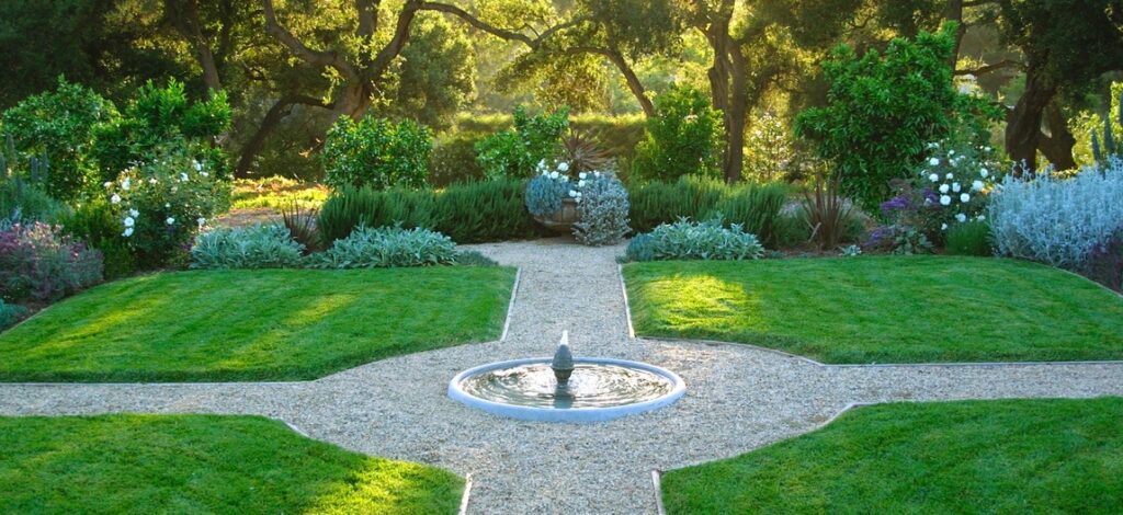 Un jardin paysagé symétrique avec une fontaine circulaire centrale, des allées en gravier divisant les pelouses verdoyantes en quatre sections, entourées de diverses plantes et arbres en arrière-plan. Ce type de conception peut être le fruit du travail d'un paysagiste ou d'une entreprise de paysagisme spécialisée dans la création et l'entretien de jardins. Une société paysagère pourrait offrir une prestation paysagère complète pour réaliser un tel espace harmonieux et bien structuré.