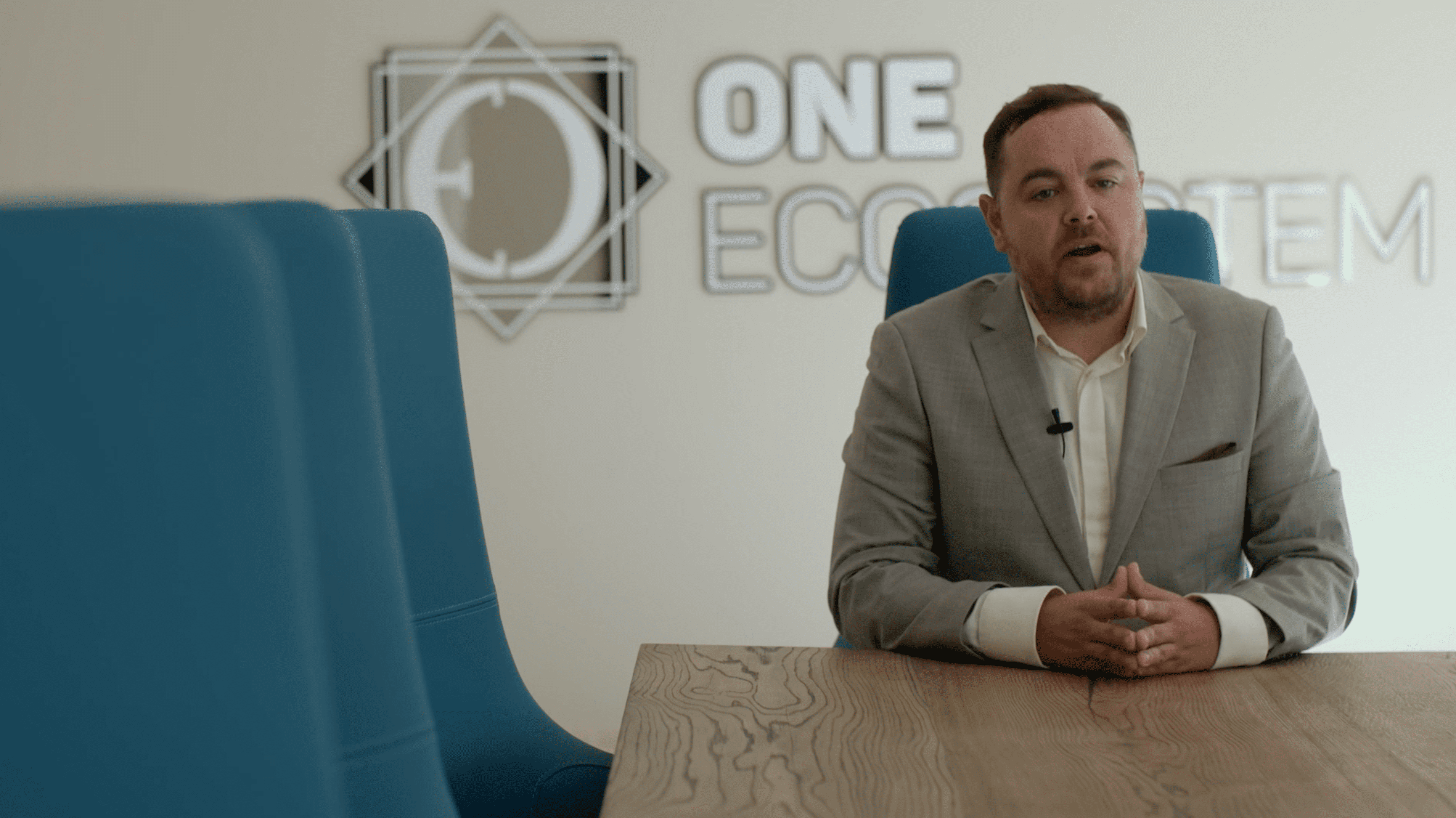 グレーのスーツを着た男性が、「ONE ECOSYSTEM」と書かれた壁の前で青い椅子のある木製の会議テーブルに座り、自社の新製品について話し合うデジタルイベントの準備をしている。