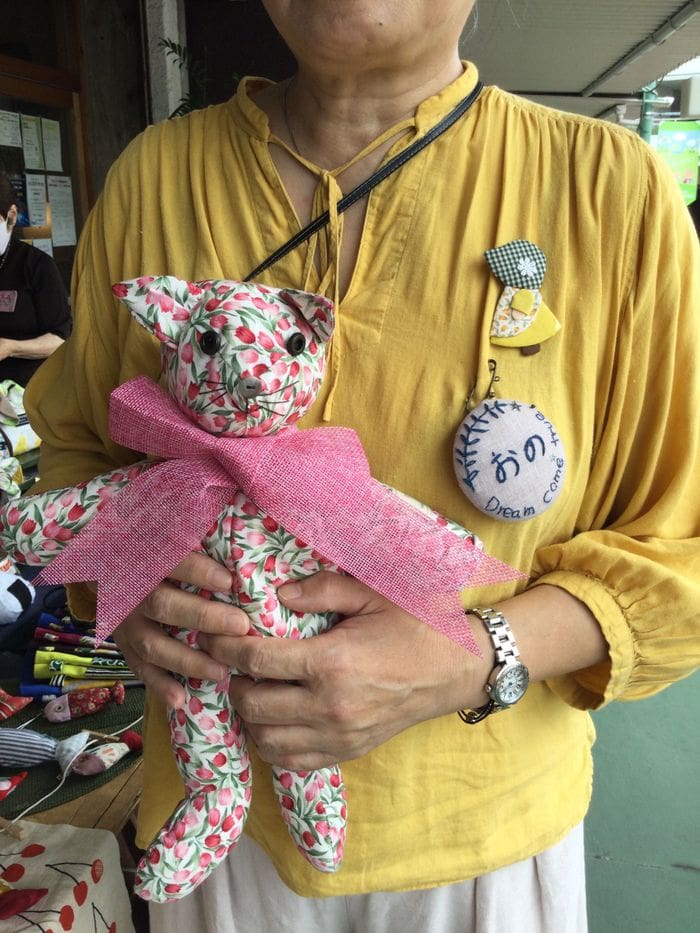 Mulher de camisa amarela segurando um gato artesanal e peixe de pelúcia.