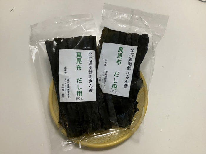 小さな黄色いザルの上に乾燥昆布の密封パックが 2 つあります。日本語でラベルが貼られたパッケージは、道南三大銘柄昆布の最高品質を示しています。
