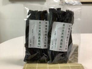 Two packs of dried kelp labeled in Japanese are laid out on a bamboo mat. One pack contains 100g of Hokkaido Eyama natural kelp. It is a set of 2 bags that reliably delivers the finest natural kelp.