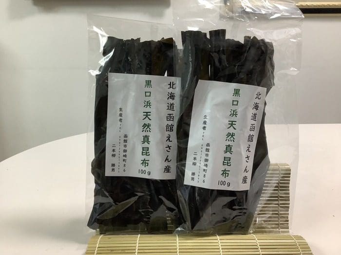 Two packs of dried kelp labeled in Japanese are laid out on a bamboo mat. One pack contains 100g of Hokkaido Eyama natural kelp. It is a set of 2 bags that reliably delivers the finest natural kelp.