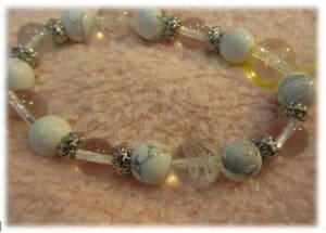 Uma pulseira de contas é colocada em uma superfície rosa suave com uma combinação de contas brancas, contas de magnesita, contas transparentes e espaçadores de prata.