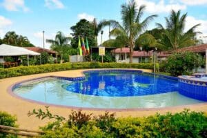 Outdoor swimming pool with crystal clear blue water, surrounded by lush greenery and palm trees. Under a cloudy sky, several small buildings with red tiled roofs can be seen in the background, making it the perfect place to stay in Quindio during an unforgettable trip to Colombia.