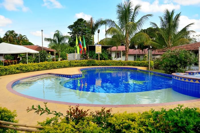 Outdoor swimming pool with crystal clear blue water, surrounded by lush greenery and palm trees. Under a cloudy sky, several small buildings with red tiled roofs can be seen in the background, making it the perfect place to stay in Quindio during an unforgettable trip to Colombia.