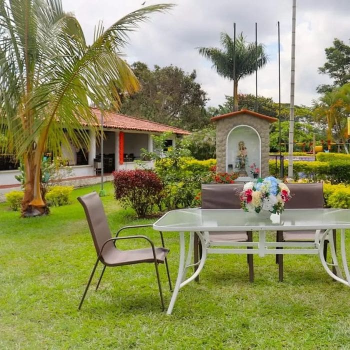 Outdoor scene with glass-top table and chairs on the lawn. Nearby, a shrine of small statues adds charm, and in the background stands a red-roofed building surrounded by palm trees. Perfect for the Campestre experience during your trip to Colombia.