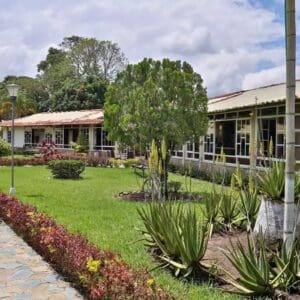 Under a slightly cloudy sky, in front of a building with white window frames, a garden planted with a variety of plants and shrubs provides a serene backdrop for your next trip to Colombia.