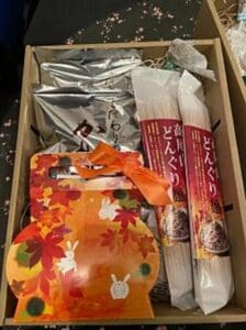 日本語ラベルの付いた食品2パック、紅葉と白うさぎが描かれたオレンジ色の装飾バッグ、ヘルシーなどんぐり商品、その他のパッケージ商品が入ったギフトボックス。