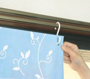 白い花柄の青いカーテンを、金属棒に取り付けられた白いフックに引っ掛けている手。この断熱カーテンは、インテリアに魅力を加えるだけでなく、エネルギーの節約にも役立ちます。