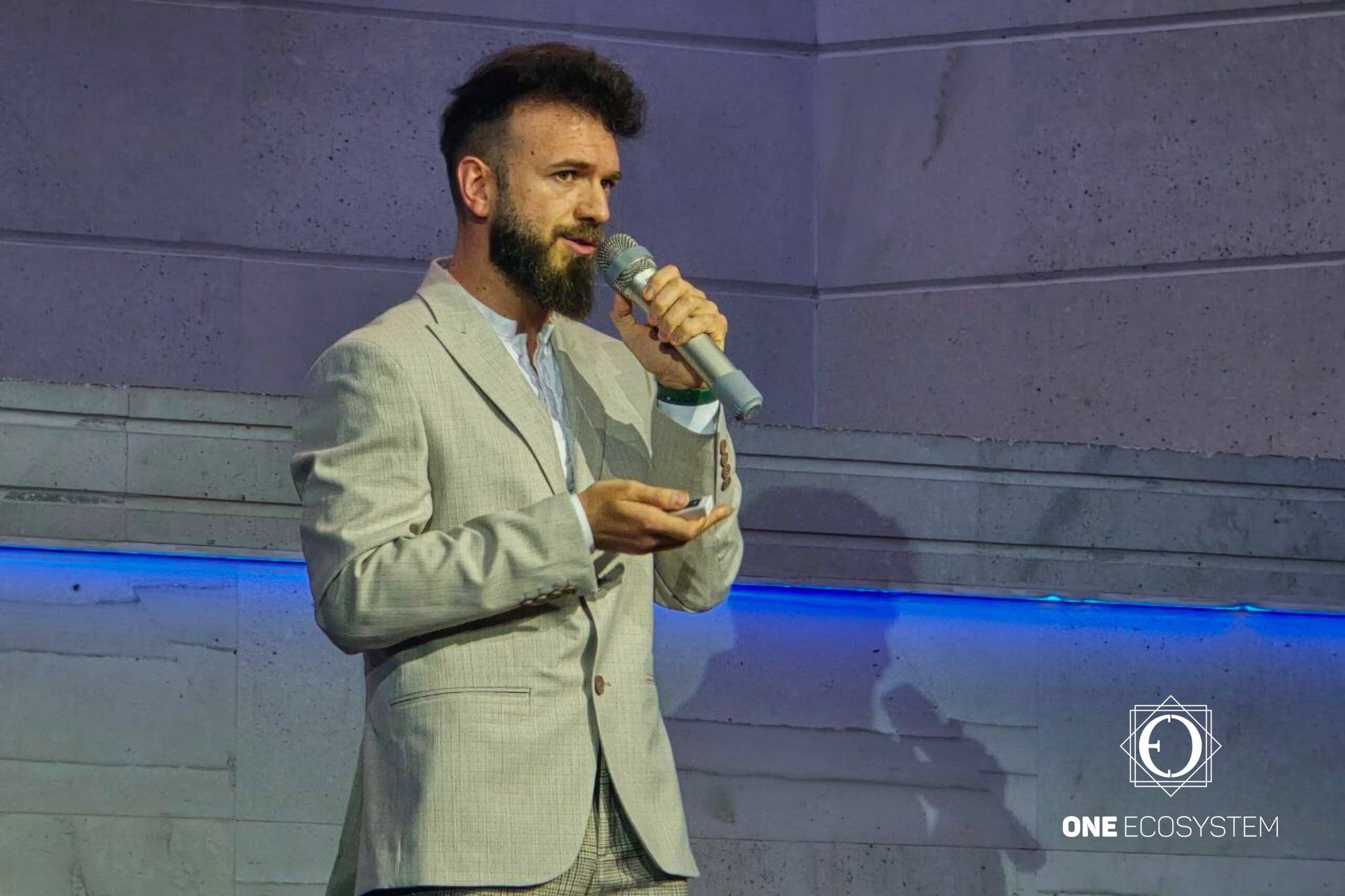 A bearded, well-trimmed man is talking into a microphone while holding a small device against the backdrop of a stone wall with blue lines and a logo that says "ONE ECOSYSTEM" in the corner. Prominent Ivan Zerkov provides insights related to guest purchases and merchants.