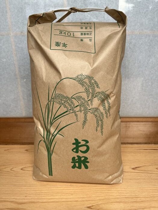 On the wooden floor, a brown paper bag containing 10 kg of fresh rice with an illustration of rice is placed. The package is printed with Japanese and the words "Shinanomachi Koshihikari Rice".