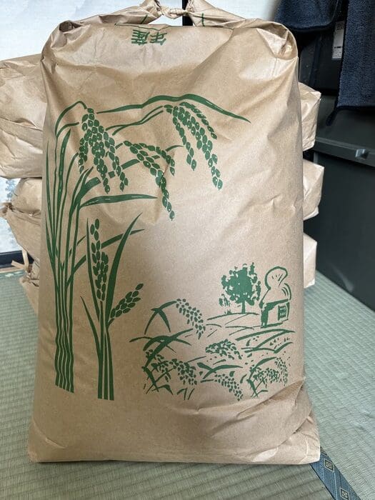 A large brown paper bag with a green illustration of rice and fields is filled with Shinanomachi Koshihikari rice, and similar bags are placed on tatami mats stacked on top of each other.
