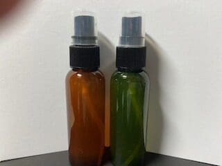 黒いキャップと透明な蓋が付いた 2 つのスプレー ボトル (1 つは茶色、もう 1 つは緑色) が白い背景に対して並べて配置されています。リラックス効果で知られる天然アロマオイルを配合したボトルです。