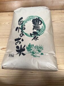 A 10kg bag of Shinanomachi Koshihikari rice with Japanese characters and green illustrations on the package is placed on a wooden surface.