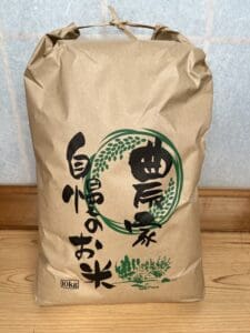 A large brown paper bag with Japanese characters on it sits upright on the wooden floor. The bag labeled 10 kg and new white rice seems to contain rice.