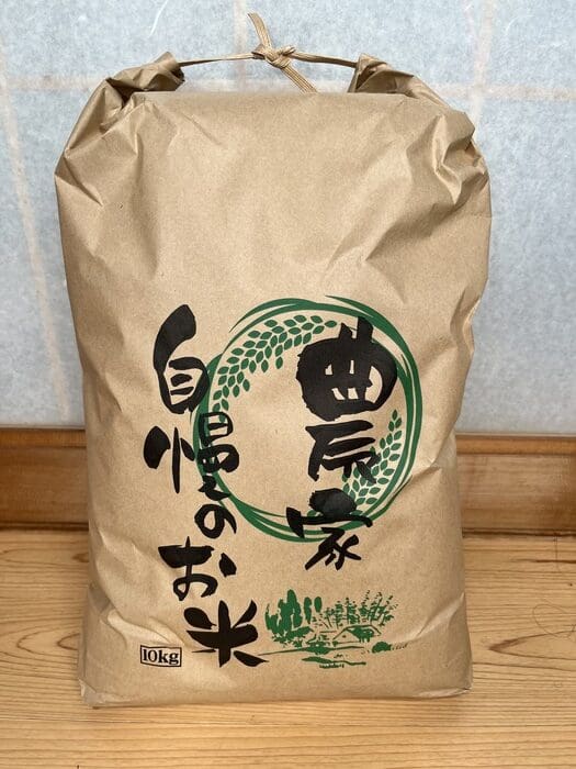 A large brown paper bag with Japanese characters on it sits upright on the wooden floor. The bag labeled 10 kg and new white rice seems to contain rice.