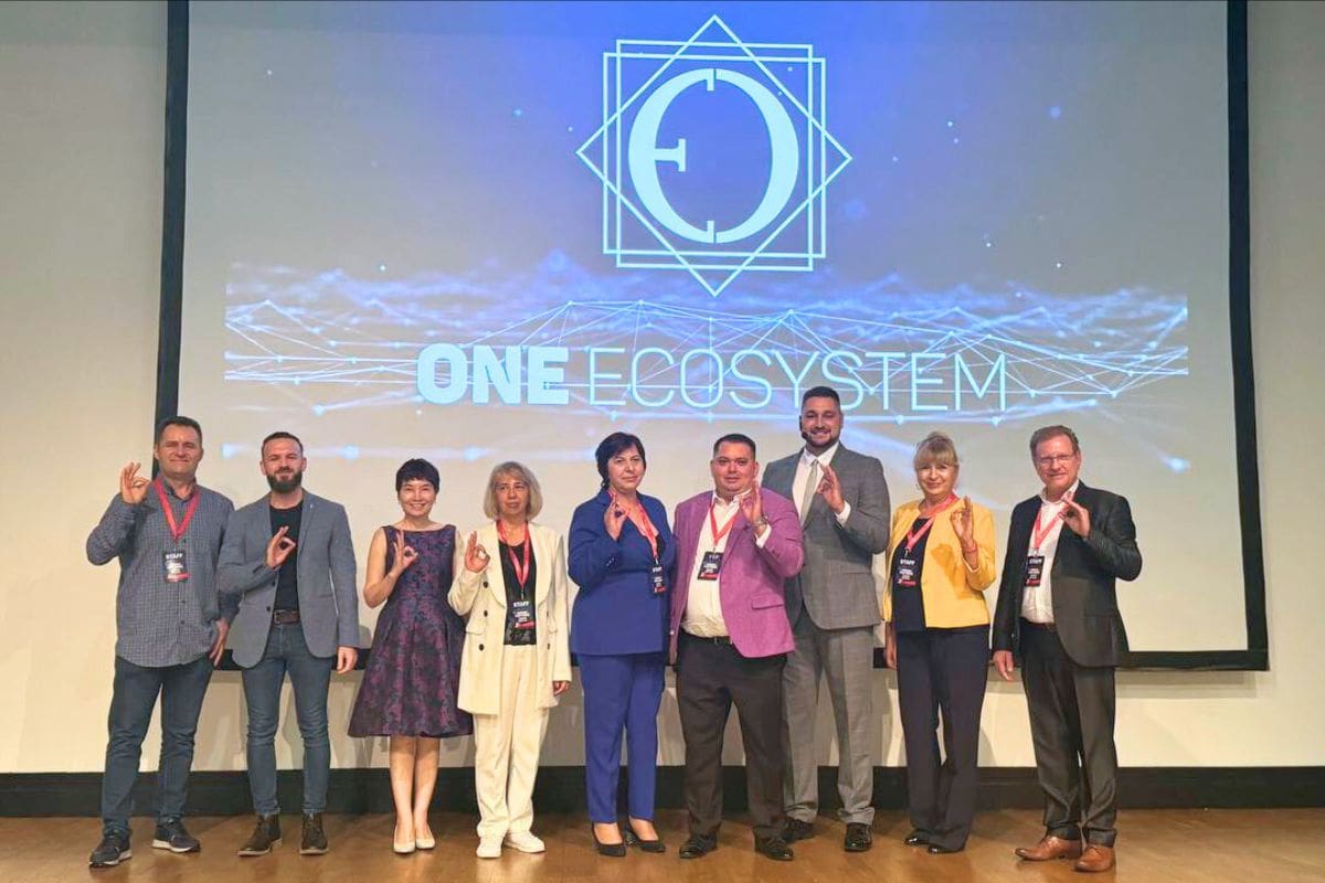Nine people are lined up on the stage, with the "One Ecosystem" logo shining in the background, each waving. The scene is part of the IMA event, which unveils exciting new features, conveying Romania's innovative spirit to a global audience.