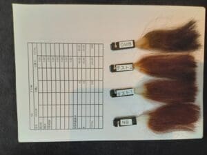Four hair samples labeled "OMB", "Test 2", "Test 1" and "NB" are placed on a table next to a datasheet with embedded rows of numbers, which are used to test various hair dye products.