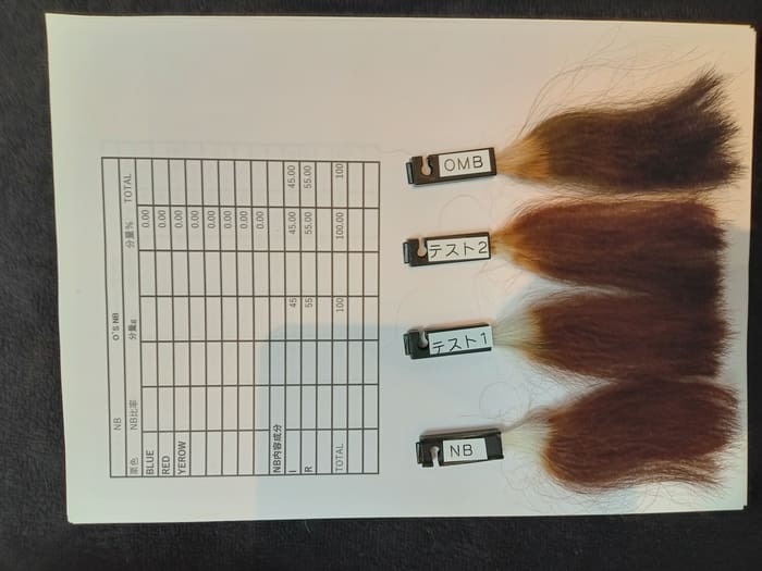 Four hair samples labeled "OMB", "Test 2", "Test 1" and "NB" are placed on a table next to a datasheet with embedded rows of numbers, which are used to test various hair dye products.