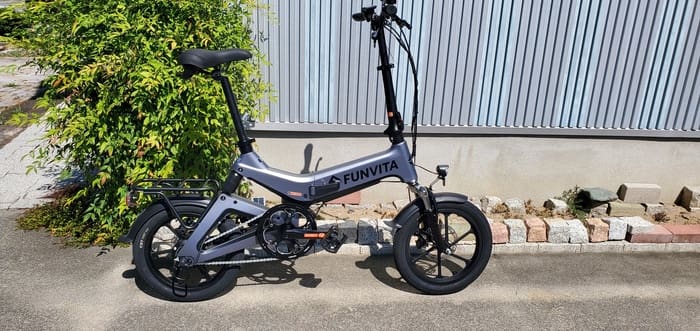 De opvouwbare elektrische fiets "FUNVITA" met 16-inch stijlvolle wielen staat geparkeerd naast een metalen hek op een betonnen weg omgeven door groen, wat een comfortabele rit belooft.