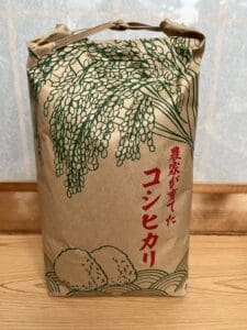 鮮やかな緑と赤の日本語で書かれたラベルに、稲と米の絵が描かれた茶色の紙袋に入ったコシヒカリ。この特別栽培米は、一粒一粒の味が保証された品質です。