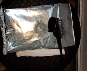 A set of supplies with a foil pack, a black bristle dye brush, and a white towel are cleverly placed on a dark circular surface.