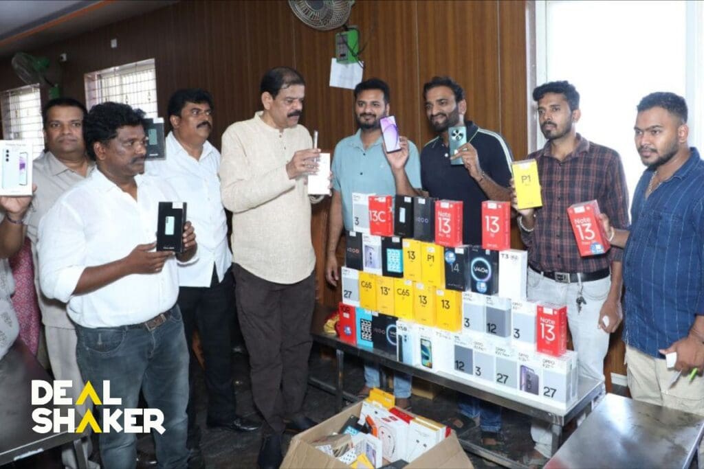 At an IMA event in Davanagelle, a group of people stood around a table lined with boxes of various smartphones in a room with wooden walls and windows.