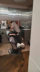 Ang isang tao na nakaupo sa isang recliner sa isang salon ay tumatanggap ng isang marangyang paghuhugas ng buhok na may henna mula sa isang estilista. Nagtatampok ang kuwartong ito ng modernong kagandahan at nagtatampok ng mga metal cabinetry at tiled floor.