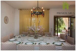 We have a restaurant with a large round table that can accommodate up to 14 people. It features a floral centerpiece and a patterned chair, with a chandelier hanging above. Opening the door leads to the garden outside, perfect for enjoying during the long weekend.