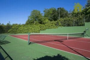 The red and green tennis courts, which can accommodate up to 14 players, are surrounded by trees and have green walls and benches on the left.