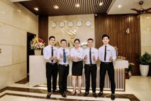 白いシャツとネクタイ姿のホテルスタッフ5名が世界時計と花飾りが飾られたフロントデスクの前に誇らしげに微笑みながら並び、思い出に残る滞在をお約束する最高の眺望とともにホテルのデラックスルームへご案内いたします。