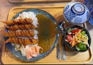 A plate of breaded shrimp on top of rice with curry sauce, red ginger and salad. Soup for side dishes with a spoon.