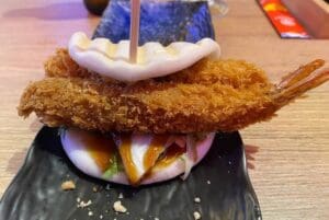 A bao ban sandwich with fried shrimp, lettuce and sauce rests on a black plate on a wooden table.