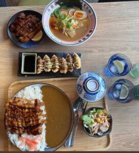 Aerial view of tables with Japan curry rice, ramen, gyoza, salad and grilled meat. Two glasses garnished with lemon slices.