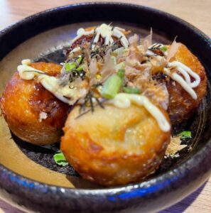 Takoyaki is served on a browned ceramic plate topped with mayonnaise, bonito flakes, and chopped green onions.