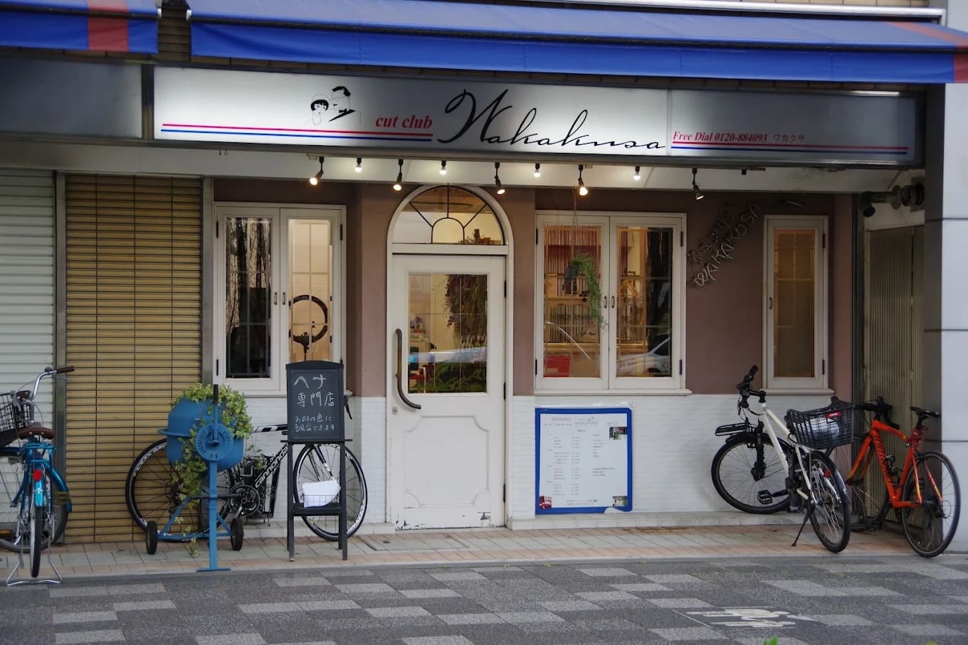 白い二重扉の上に「cut club Wakana」と書かれた看板のある店構えで、ショートカットとヘナのサービスを提供している。窓と駐輪場に挟まれ、青い自転車ラック、観葉植物、入り口付近にはメニューや看板がある。.