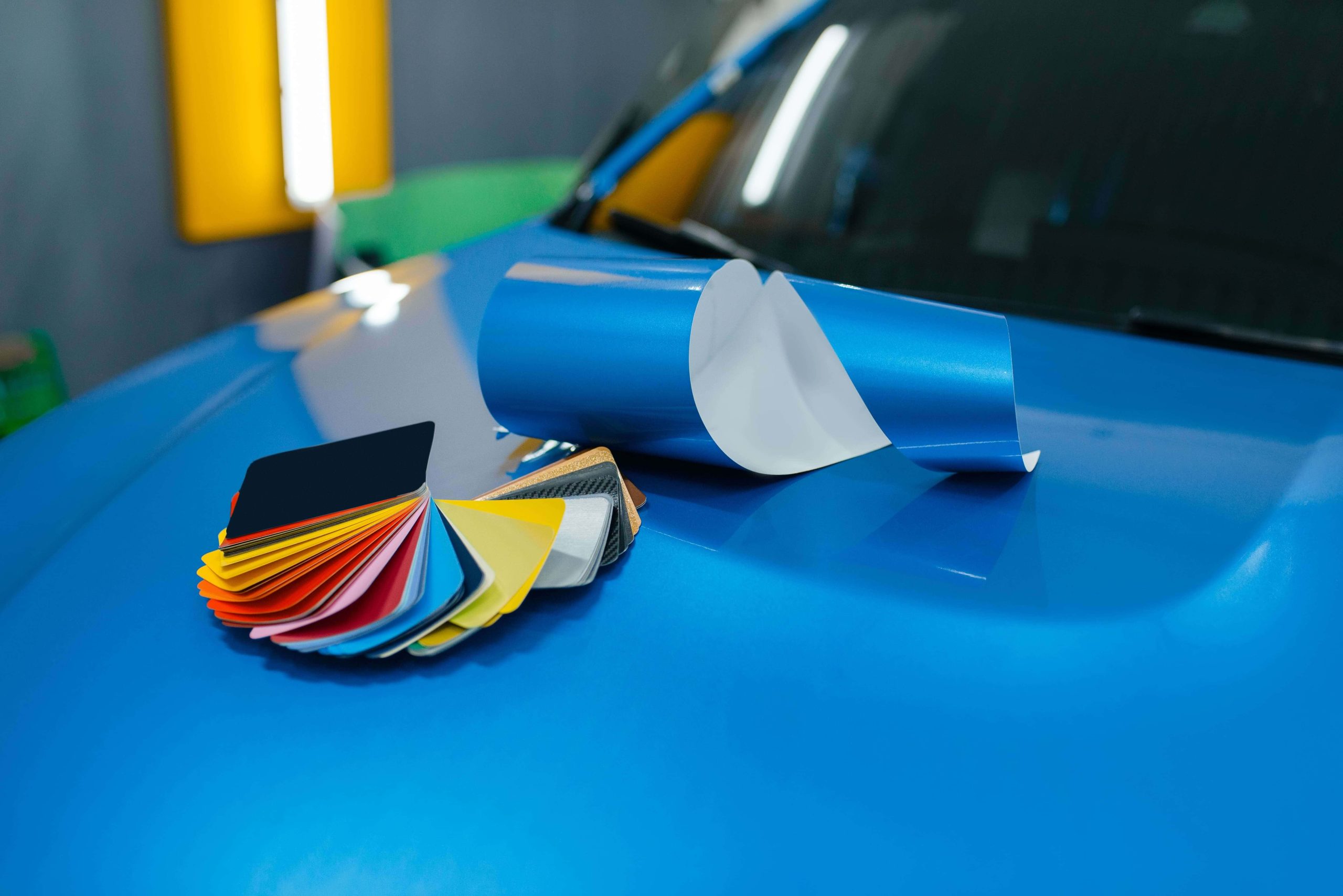 Rollo de vinilo azul y muestrario de colores sobre el capó de un coche, listos para aplicar en rotulación.